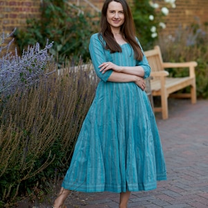 May include: A woman wearing a blue and white striped dress with a long skirt and three-quarter sleeves. She is standing on a brick walkway with a brick wall and a bench in the background.