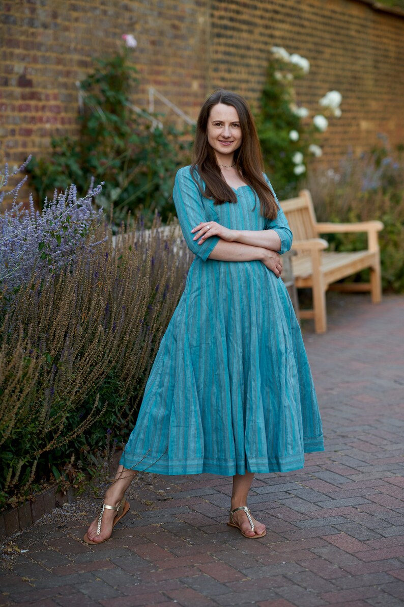 May include: A woman wearing a blue and white striped dress with a long skirt and three-quarter sleeves. She is standing on a brick walkway with a brick wall and a bench in the background.
