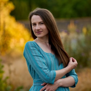 May include: A woman with long brown hair is wearing a blue and white striped dress. She is standing in a field with a blurred background of green trees and yellow flowers.