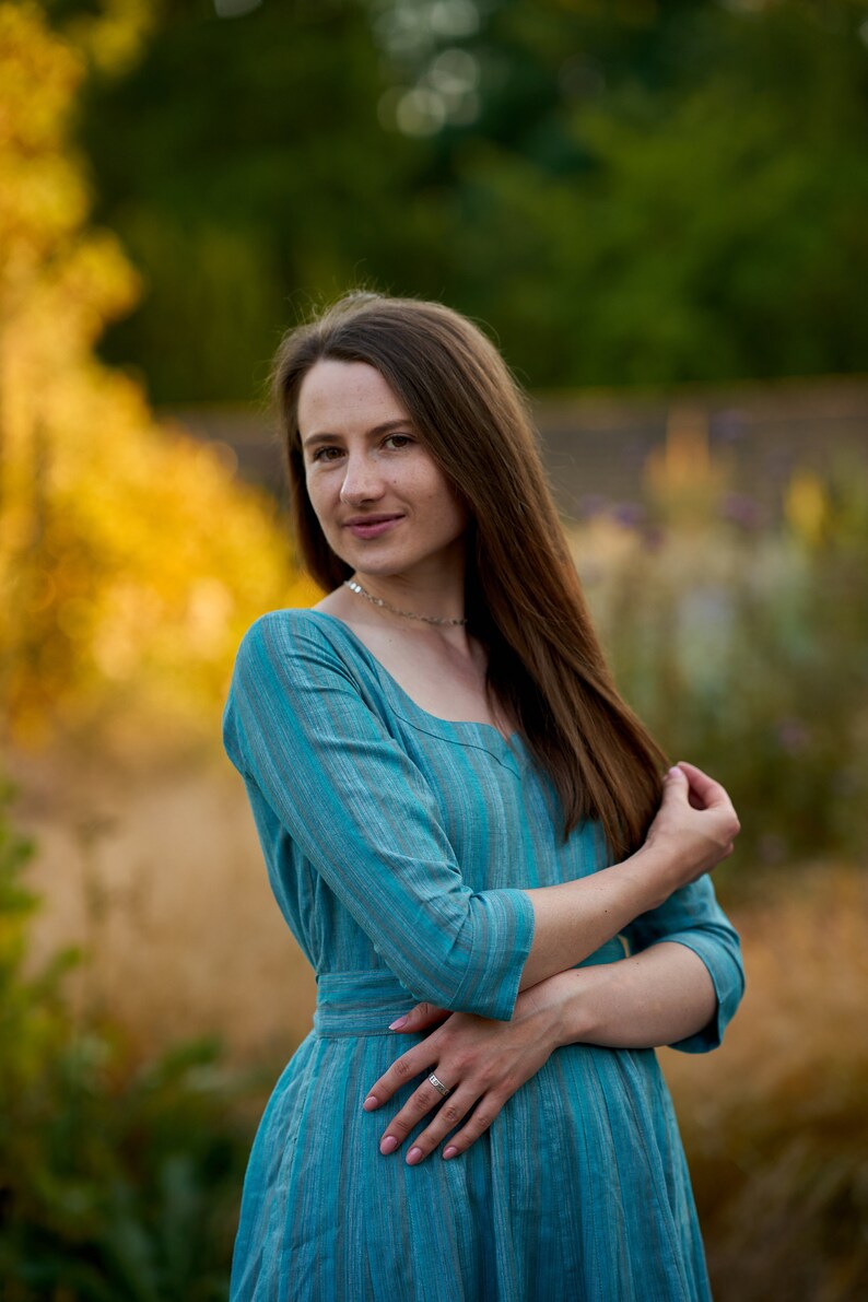 May include: A woman with long brown hair is wearing a blue and white striped dress. She is standing in a field with a blurred background of green trees and yellow flowers.