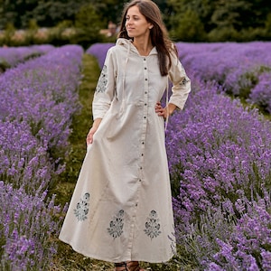 Può includere: Un lungo abito in lino beige con cappuccio e abbottonatura. L'abito presenta motivi floreali neri sulle maniche e sulla gonna. La donna è in piedi in un campo di lavanda viola.