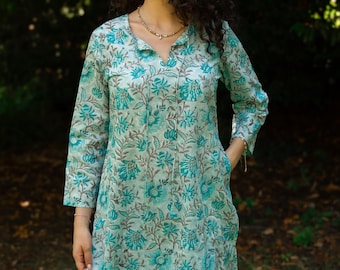 Op de afbeelding: Een lichtgroene tuniekjurk met een bloemenpatroon in teal en bruin. De jurk heeft lange mouwen, een V-hals en zakken. De vrouw draagt een ketting en een blauwe spijkerbroek. De achtergrond is een vage buitenscène.