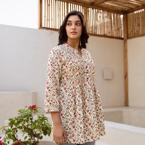 Puede incluir: Una túnica de algodón blanco con estampado floral en tonos rojos y verdes. La túnica tiene un frente abotonado y una cintura fruncida.