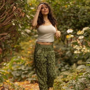 Peut inclure: Une personne porte un débardeur gris clair et un pantalon vert olive à motifs, marchant sur un sentier entouré de verdure. Le pantalon a un motif floral. Elle porte des sandales et un collier.