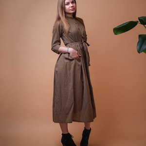 May include: A woman wearing a long, olive green dress with a button-up front and a belt. She is standing on a light brown background and has her hand in her pocket.