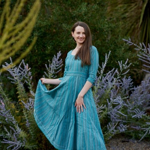 May include: A woman wearing a blue and white striped dress with a square neckline and 3/4 sleeves. She is standing in a garden with purple flowers.