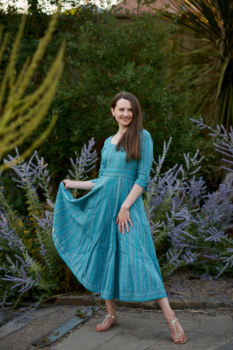 May include: A woman wearing a blue and white striped dress with a square neckline and 3/4 sleeves. She is standing in a garden with purple flowers.