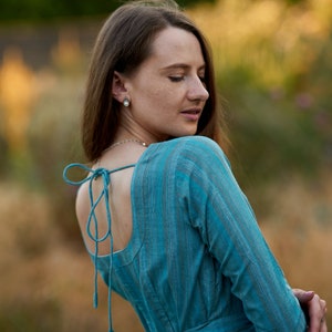 May include: A woman wearing a blue and white striped dress with a low back and a tie at the waist. The dress is made of a soft, flowing fabric.