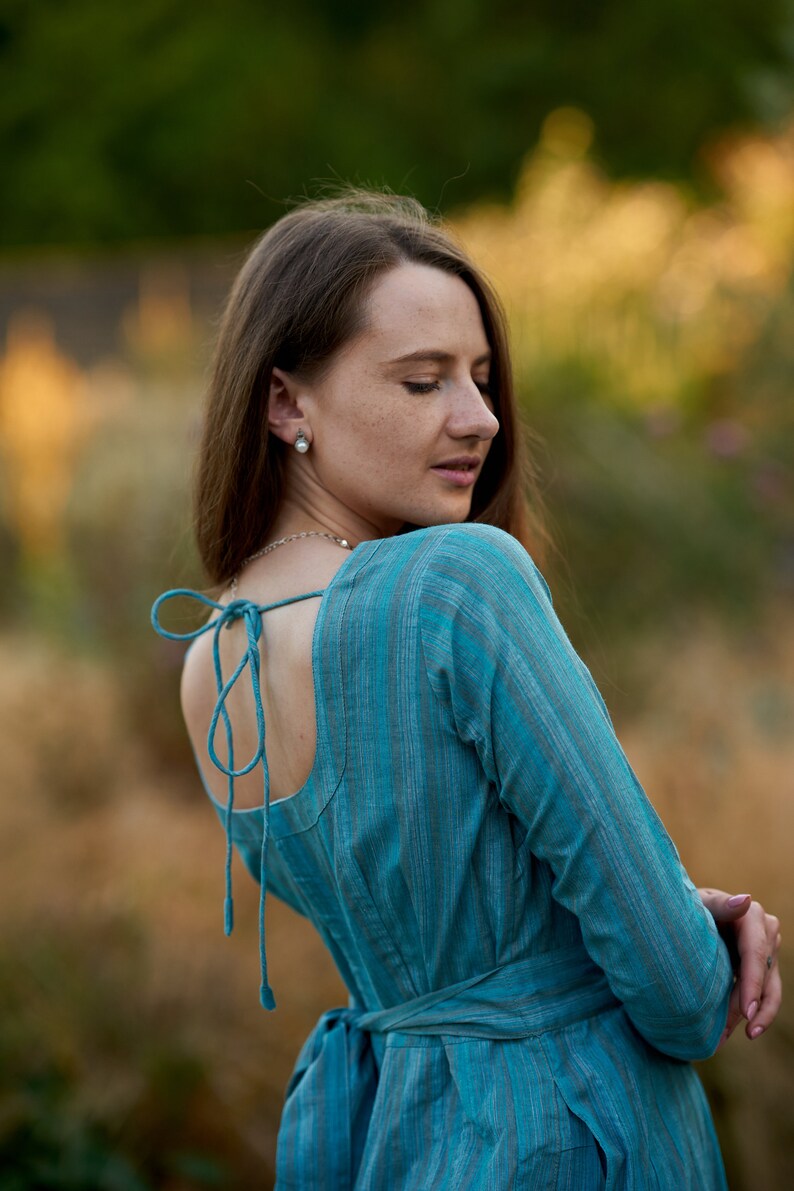 May include: A woman wearing a blue and white striped dress with a low back and a tie at the waist. The dress is made of a soft, flowing fabric.