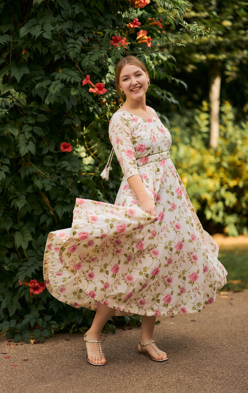 May include: A woman wearing a white floral dress with a green sash and gold sandals. She is standing in front of a green leafy wall with pink flowers.