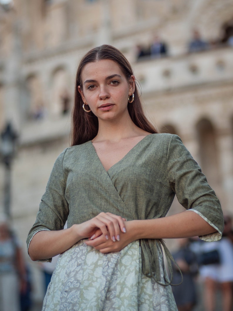 May include: A woman wearing a green linen wrap dress with a floral print underneath. The dress has a V-neckline and 3/4 sleeves.