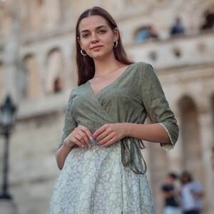 May include: A woman wearing a green wrap dress with a white floral print skirt. The dress has a V-neckline and a tie at the waist. She is standing in front of a building with a stone facade.
