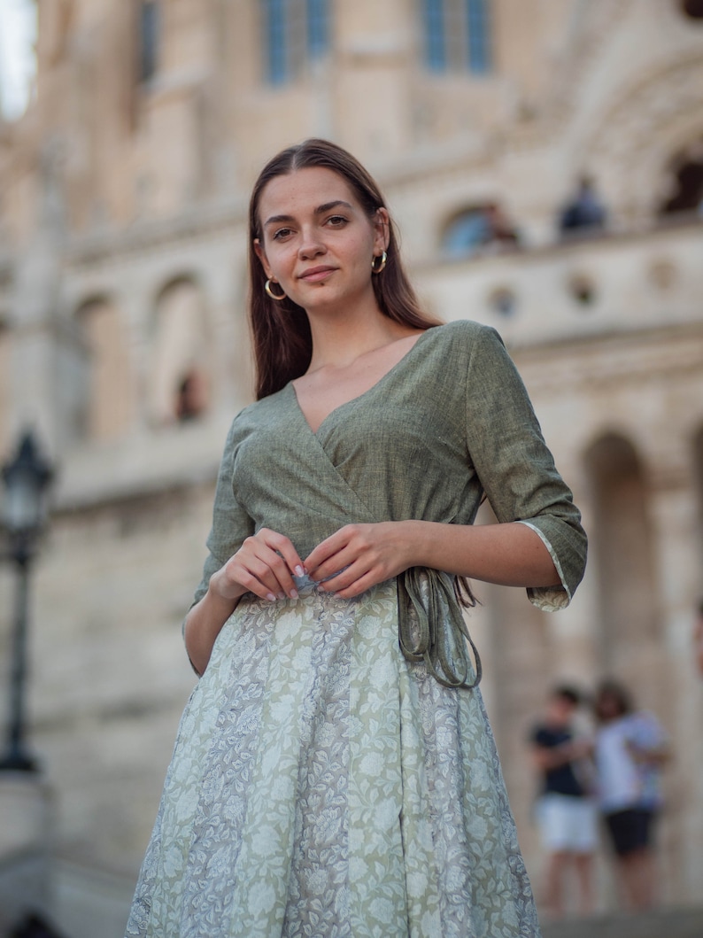 May include: A woman wearing a green wrap dress with a white floral print skirt. The dress has a V-neckline and a tie at the waist. She is standing in front of a building with a stone facade.