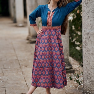 May include: A woman wearing a blue and pink patterned dress with a blue top and a red and gold trim. She is standing in a garden setting with pillars in the background.