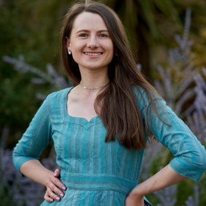 May include: A woman wearing a teal blue dress with thin white stripes. The dress has a sweetheart neckline and a fitted waist. She is standing in front of a blurred background of green foliage.