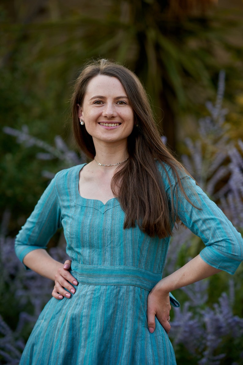 May include: A woman wearing a teal blue dress with thin white stripes. The dress has a sweetheart neckline and a fitted waist. She is standing in front of a blurred background of green foliage.