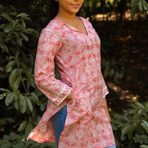 Puede incluir: Una túnica rosa con estampado floral y cuello en V. La túnica tiene mangas largas con adornos blancos y dorados en los puños. La persona lleva vaqueros azules. El fondo es un entorno verde y frondoso.