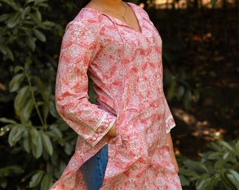 Op de afbeelding: Een roze tuniek met een bloemenpatroon en een V-hals. De tuniek heeft lange mouwen met wit en goudkleurige afwerking aan de manchetten. De persoon draagt een blauwe spijkerbroek. De achtergrond is een groene, bladerrijke omgeving.