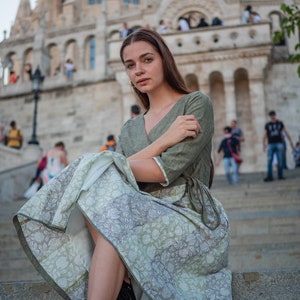 May include: A woman wearing a green linen dress with a floral print skirt sits on stone steps in front of a large stone building.