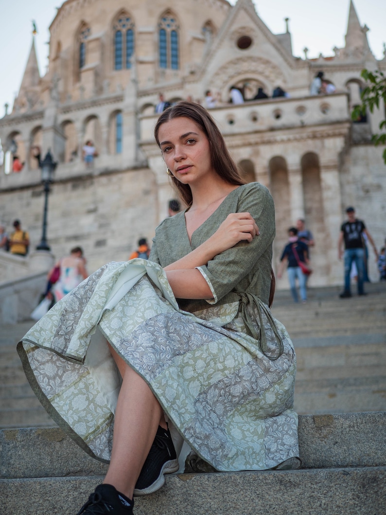 May include: A woman wearing a green linen dress with a floral print skirt sits on stone steps in front of a large stone building.