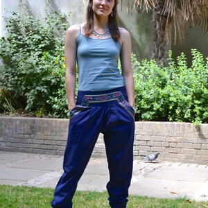 May include: A woman wearing a blue tank top and blue pants with a colorful embroidered waistband. She is standing barefoot on a grassy lawn.