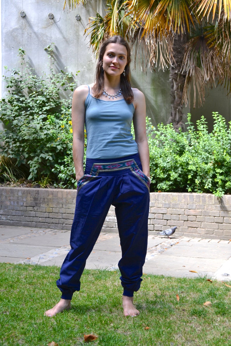 May include: A woman wearing a blue tank top and blue pants with a colorful embroidered waistband. She is standing barefoot on a grassy lawn.