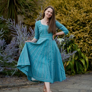 May include: A woman wearing a blue and white striped dress with a white petticoat underneath. She is standing on a stone path with green plants in the background.