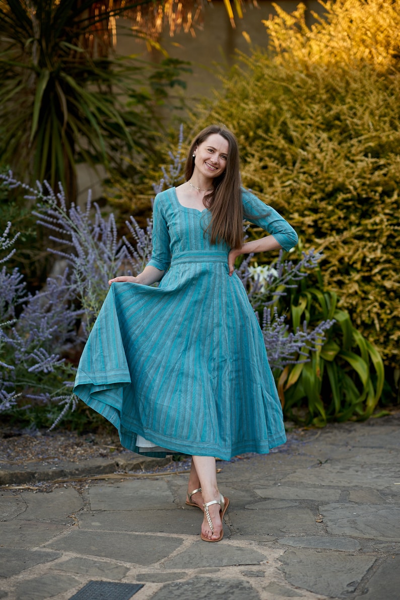 May include: A woman wearing a blue and white striped dress with a white petticoat underneath. She is standing on a stone path with green plants in the background.