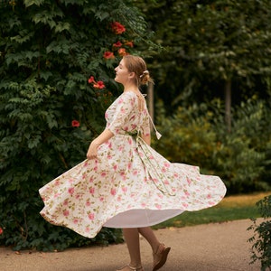 May include: A woman in a white floral dress with a green sash spins around in a park. The dress has a white petticoat underneath and the woman is wearing gold sandals.
