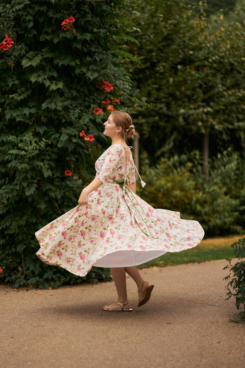 May include: A woman in a white floral dress with a green sash spins around in a park. The dress has a white petticoat underneath and the woman is wearing gold sandals.