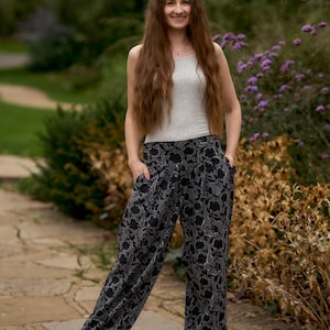 Könnte beinhalten: Eine Frau trägt ein graues Tanktop und eine schwarze und weiße Hose mit Blumenmuster. Sie steht auf einem Steinweg mit grünem Laub und lila Blumen im Hintergrund.