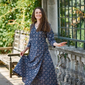 Könnte beinhalten: Eine Frau in einem blau-weiß gemusterten Kleid mit V-Ausschnitt und 3/4-Ärmeln. Sie steht vor einer Steinmauer mit einem Metallgitter dahinter.