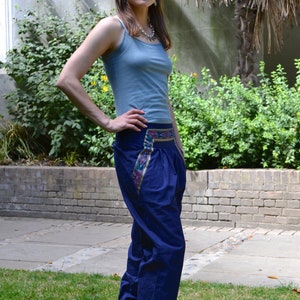May include: A woman wearing a light blue tank top and dark blue pants with a colorful embroidered waistband. She is standing on a grassy lawn.