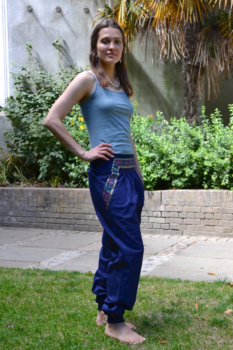 May include: A woman wearing a light blue tank top and dark blue pants with a colorful embroidered waistband. She is standing on a grassy lawn.