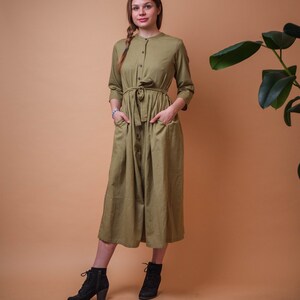 Könnte beinhalten: Eine Frau trägt ein langes, olivgrünes, knopfloses Kleid mit einem Bindegürtel. Sie steht vor einem hellbraunen Hintergrund und hat die Hände in den Taschen.