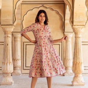 May include: A woman wearing a beige wrap dress with a pink floral print. The dress has a V-neckline and 3/4 sleeves. She is standing in a hallway with arched doorways.