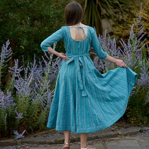 May include: A woman wearing a blue and white striped dress with a low back and a tie at the waist. She is standing in a garden with purple flowers.