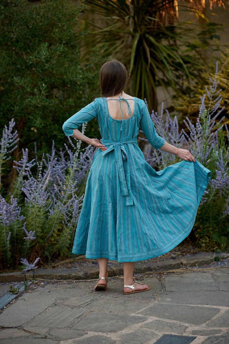 May include: A woman wearing a blue and white striped dress with a low back and a tie at the waist. She is standing in a garden with purple flowers.