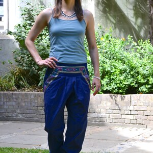 May include: A woman wearing a blue tank top and blue pants with a colorful patterned waistband. She is standing in a grassy area with a brick wall and greenery in the background.