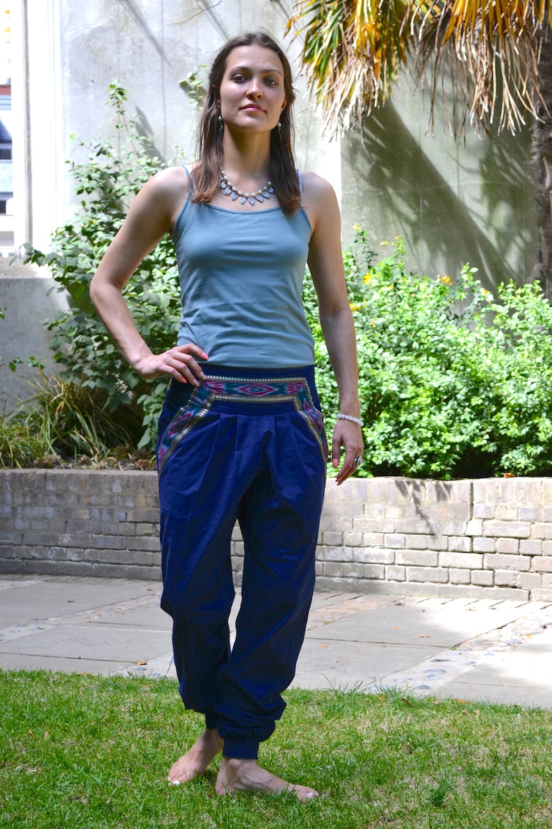 May include: A woman wearing a blue tank top and blue pants with a colorful patterned waistband. She is standing in a grassy area with a brick wall and greenery in the background.