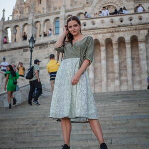 May include: A woman wearing a green and white patterned dress stands on a set of stone steps in front of a large building.
