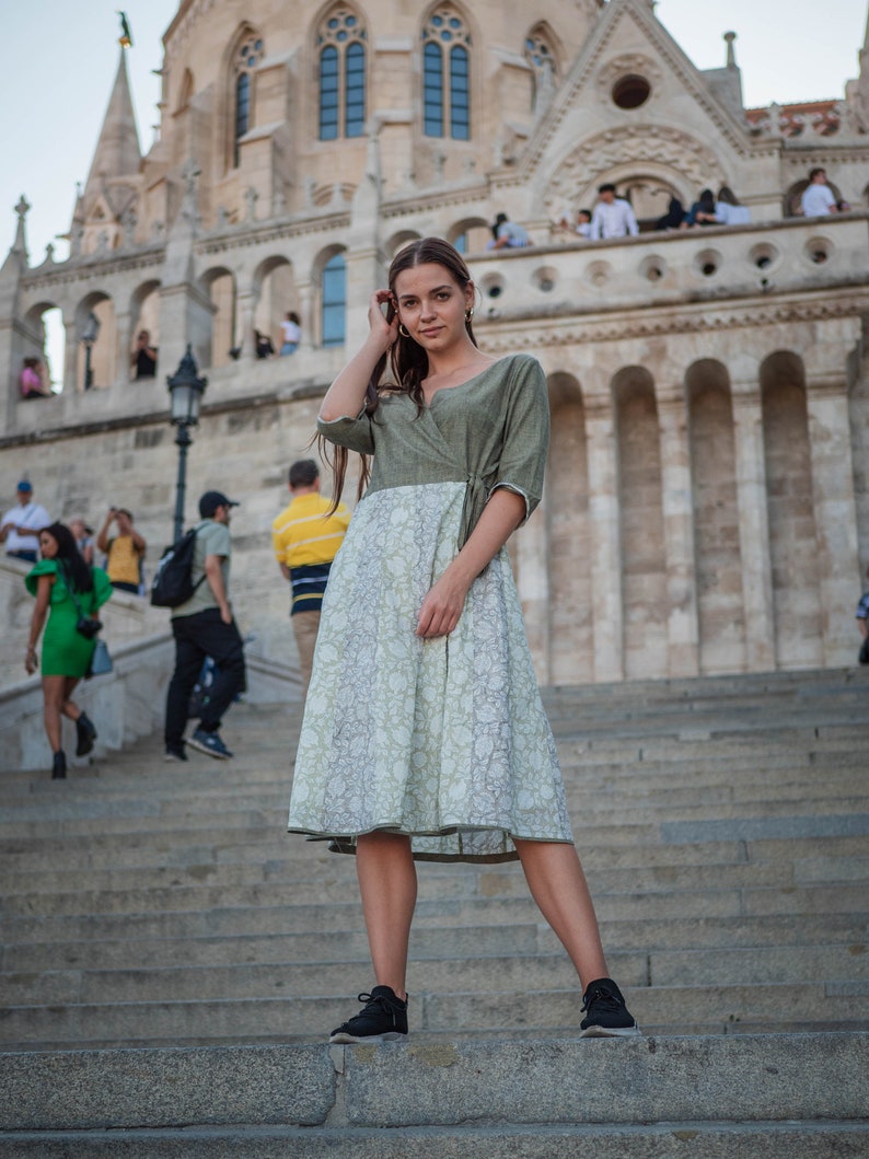 May include: A woman wearing a green and white patterned dress stands on a set of stone steps in front of a large building.