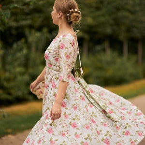 May include: A woman wearing a white dress with a pink floral pattern and a green sash. The dress has a scoop neckline and 3/4 sleeves. The woman is standing in a park with green trees in the background.