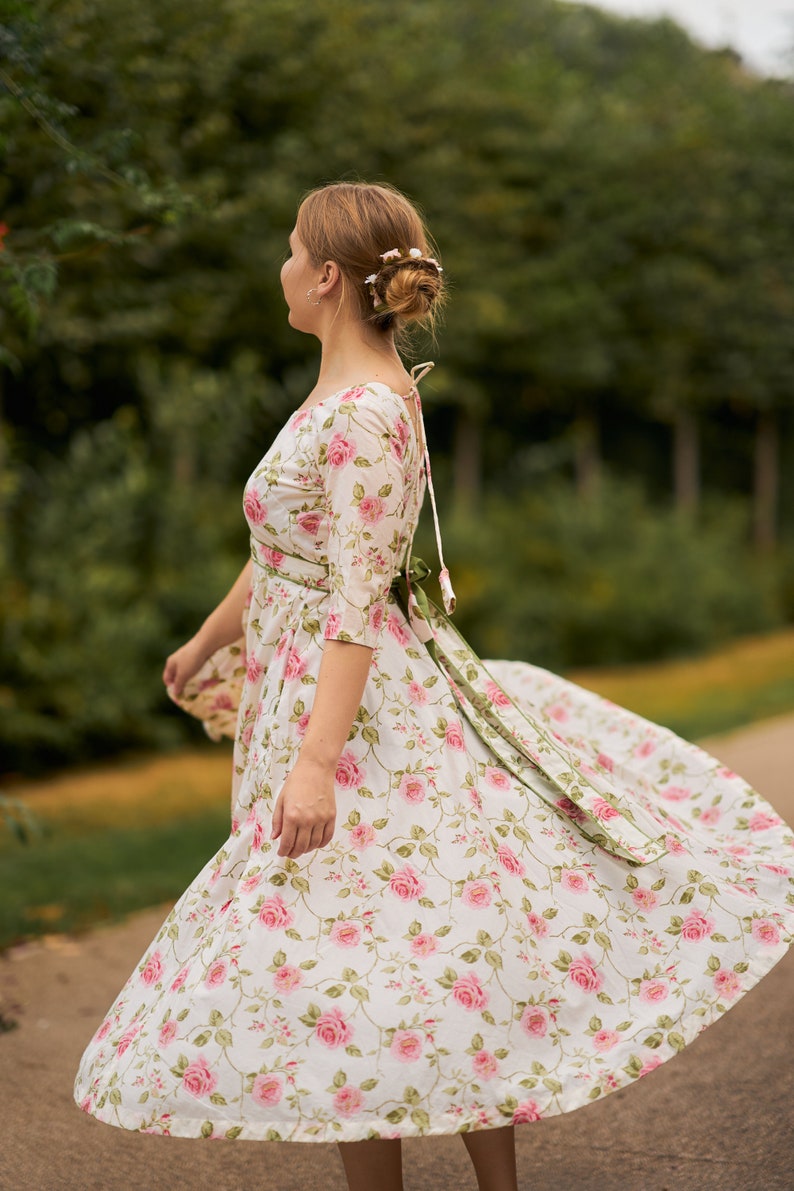 May include: A woman wearing a white dress with a pink floral pattern and a green sash. The dress has a scoop neckline and 3/4 sleeves. The woman is standing in a park with green trees in the background.