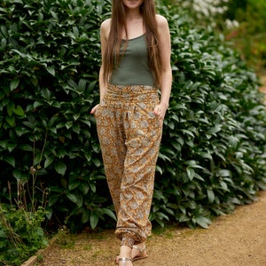 May include: A woman wearing a green tank top and patterned trousers stands on a path in a garden. She is smiling and looking at the camera.