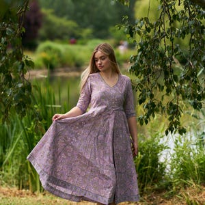 May include: A light purple wrap dress with a floral pattern and elbow-length sleeves. The dress has a V-neck and a flowing skirt. The model is wearing brown sandals. The background is a park with a lake and trees.