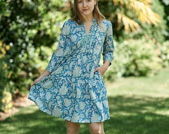 Op de afbeelding: Een vrouw draagt een blauwe en witte bloemenjurk met een groene bies. Ze staat in een grasachtig gebied met bomen op de achtergrond.