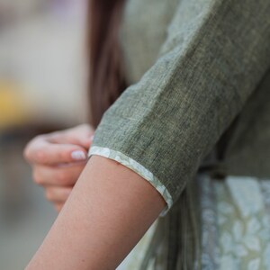 May include: A close-up of a green linen shirt sleeve with a white floral trim. The sleeve is rolled up to the elbow, showing the fabric's texture.
