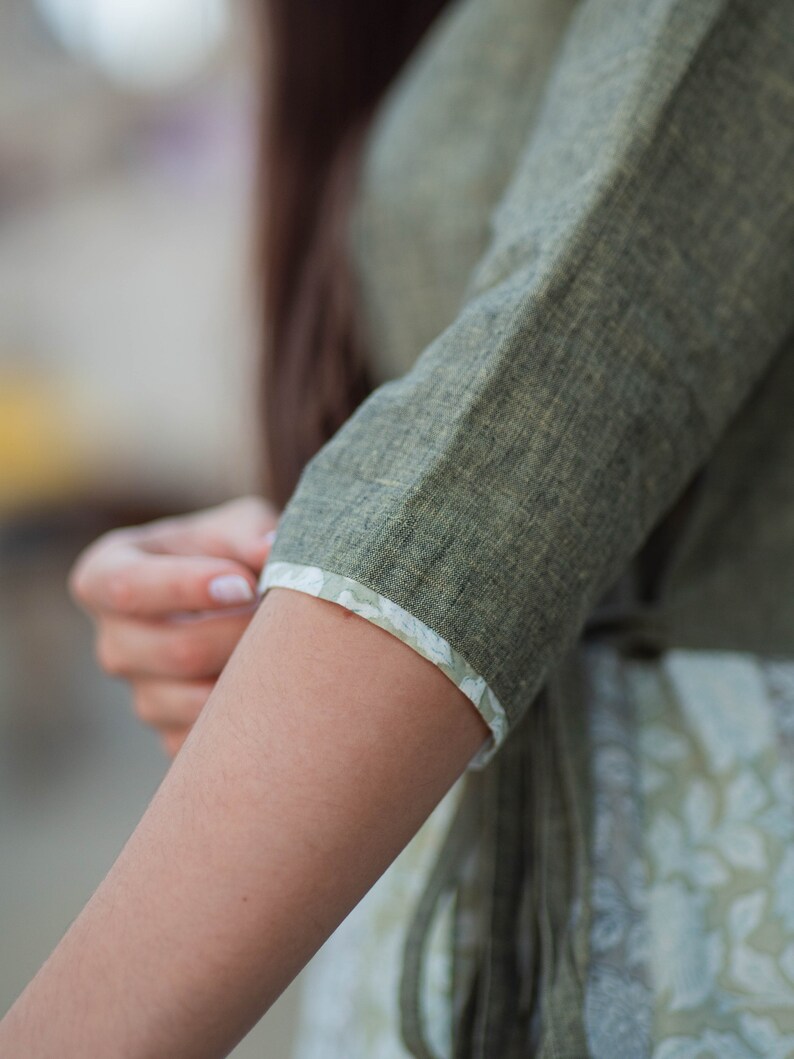May include: A close-up of a green linen shirt sleeve with a white floral trim. The sleeve is rolled up to the elbow, showing the fabric's texture.