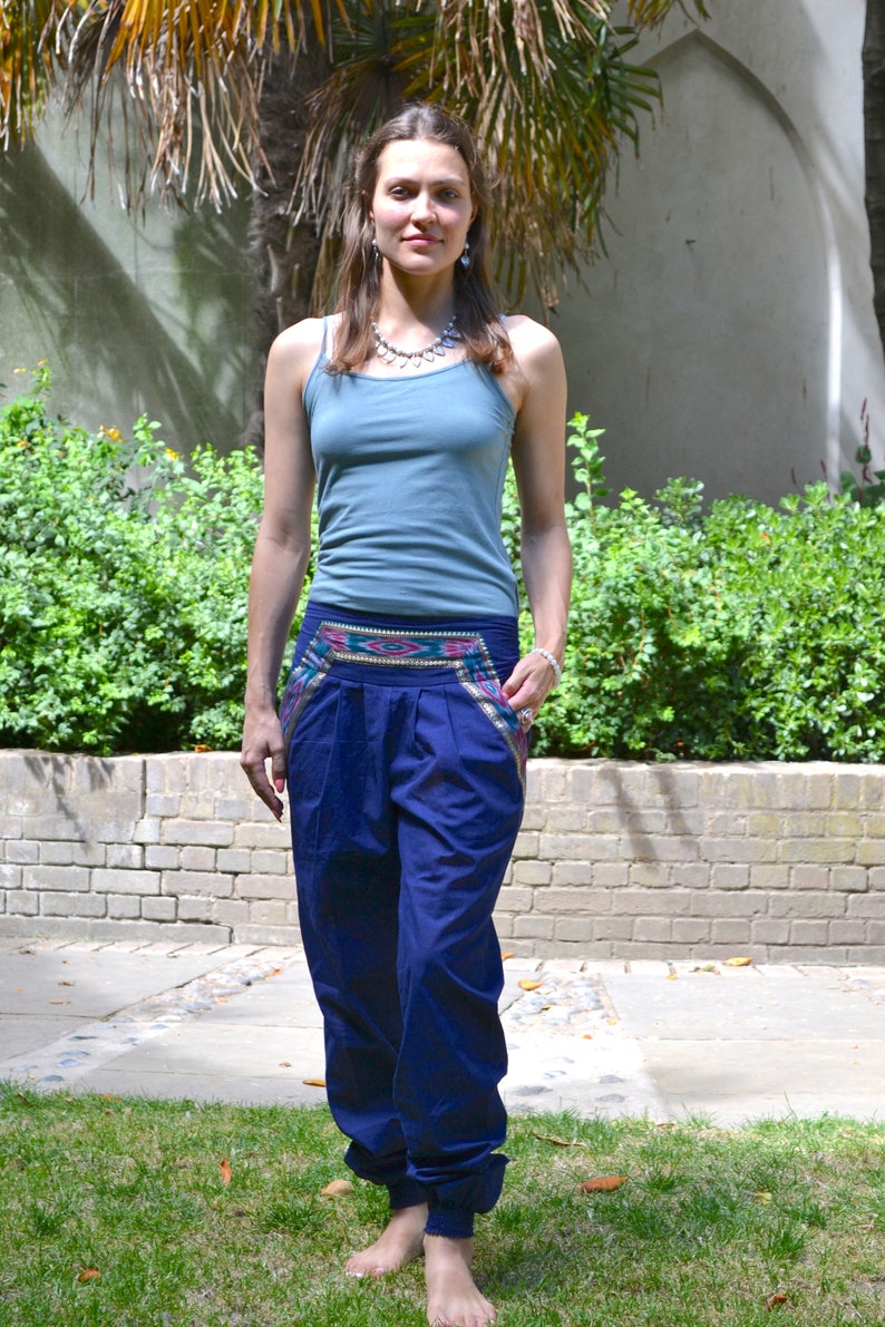 May include: A woman wearing a blue tank top and dark blue pants with a colorful patterned waistband. She is standing in front of a brick wall and green plants.